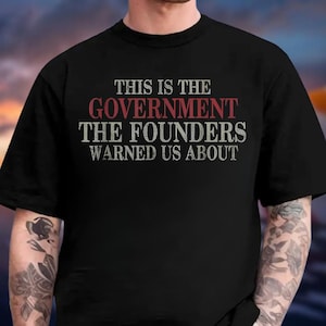 May include: Black t-shirt with the text "THIS IS THE GOVERNMENT THE FOUNDERS WARNED US ABOUT" in white and red lettering. The shirt has a crew neck and short sleeves.