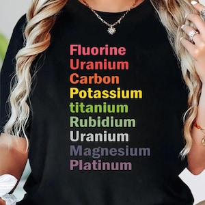 May include: A black t-shirt featuring a vertical list of chemical elements printed in various colors: Fluorine, Uranium, Carbon, Potassium, titanium, Rubidium, Uranium, Magnesium, and Platinum. The text is arranged in a column on the front center.
