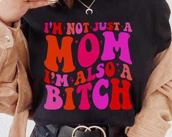May include: Black t-shirt with the text "I'M NOT JUST A MOM I'M ALSO A BITCH" in retro-style, colorful lettering. The shirt is paired with dark jeans and a black belt with a gold buckle.