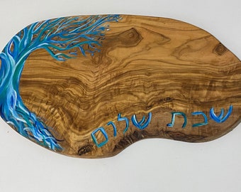May include: An olive wood serving board with a blue painted tree design on the left side. The board has the Hebrew words "Shabbat Shalom" painted in blue. The wood grain is visible.