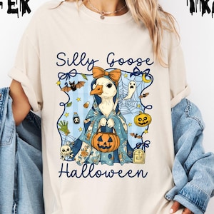 Puede incluir: Camiseta color crema con un diseño de ganso de dibujos animados para Halloween. El ganso lleva una capa azul y sostiene una calabaza. El texto "Silly Goose Halloween" está presente. Murciélagos, fantasmas y otros elementos de Halloween rodean al ganso.