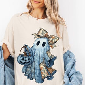 May include: Cream-colored t-shirt featuring a watercolor illustration of a friendly ghost holding a blue pumpkin bucket. The ghost has a floral bow and patchwork details. The shirt is part of a Halloween-themed collection.