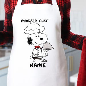 May include: White apron with a cartoon chef design featuring Snoopy, the Peanuts character, holding a tray. The apron has the words "MASTER CHEF" and "NAME" printed on it. A red mug with white hearts and a red kitchen towel are also visible.