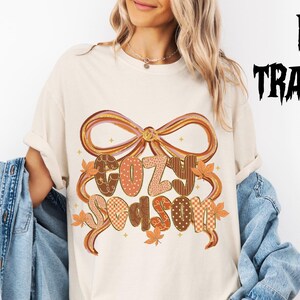 Puede incluir: Camiseta color crema con un gráfico temático de otoño. El diseño presenta las palabras "Cozy Season" en una fuente decorativa, rodeadas por un lazo y hojas de otoño. Las palabras son en tonos marrón, naranja y crema. La imagen también incluye el texto "DTF TRANSFER" en negro.