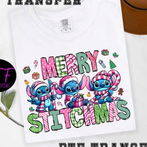 May include: White t-shirt with a festive graphic of three blue cartoon characters in Santa hats and scarves, holding candy canes and gifts. The words "MERRY STITCHMAS" are written in colourful, patterned letters.
