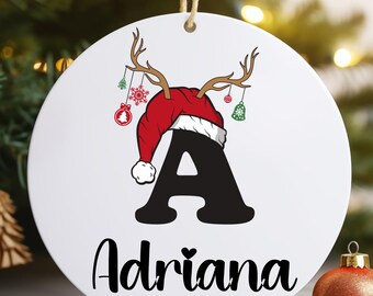 May include: White round Christmas bauble featuring a red Santa hat, reindeer antlers, and the letter A in black with the name Adriana written below.