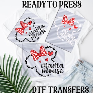 Puede incluir: Dos camisetas blancas con un diseño en blanco y negro de orejas de Mickey Mouse con el texto "mama mouse" en negro y corazones rojos. El diseño está en una hoja de transferencia transparente, lista para ser presionada sobre las camisetas. Transferencias DTF.