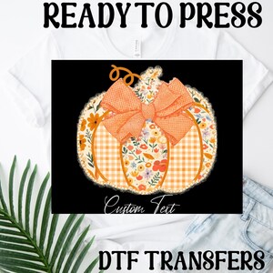 Puede incluir: Una camiseta blanca con un diseño de calabaza. La calabaza está decorada con un estampado floral, cuadros de vichy naranja y un lazo naranja. El texto "READY TO PRESS" y "DTF TRANSFERS" está arriba y abajo.