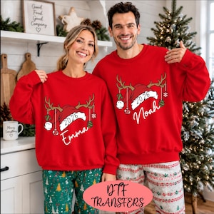 Könnte beinhalten: Zwei rote Sweatshirts mit Rundhalsausschnitt, bedruckt mit einer Weihnachtsmütze, Rentiergeweih, hängenden Christbaumkugeln, der Jahreszahl 2026 sowie den Namen Emma und Noah in weißer Schrift. Die Sweatshirts haben lange Ärmel und einen Rippkragen.