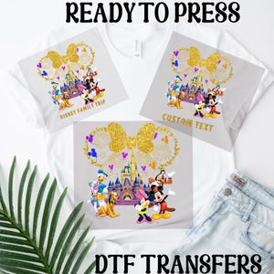 Puede incluir: Dos camisetas blancas con transferencias de planchado que muestran un lazo dorado, fuegos artificiales, el castillo de Cenicienta y personajes de Disney. Una camiseta dice "Disney Family Trip" y la otra dice "Custom Text". El texto "DTF Transfers" está en la parte inferior de la imagen.