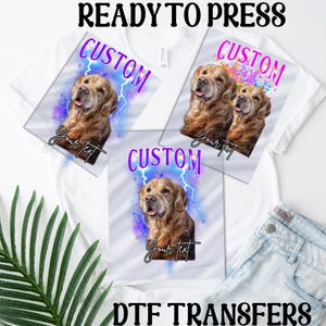 Pode incluir: Três camisetas brancas com designs personalizados que apresentam cães golden retriever. Os designs incluem um raio roxo, um fundo de tinta colorida salpicada e um fundo de aquarela roxo e azul. O texto "CUSTOM" está escrito em letras de purpurina roxas em cada camiseta. O texto "Your Text" está escrito em preto em cada camiseta. As camisetas estão prontas para serem impressas com transferências DTF.