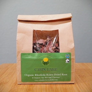 Op de afbeelding: Een bruine papieren zak met een doorzichtig venster met gedroogde Rhodiola Rosea-wortel. De zak heeft een groen label met de tekst "CABIN LAKE" en "Organic Rhodiola Rosea Dried Root, 227 gram". De zak staat op een houten oppervlak.