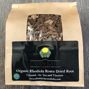 May include: A brown paper bag with a clear window showing dried Rhodiola Rosea root. The bag has a black label with the text "CABIN LAKE FARMS" and "Organic Rhodiola Rosea Dried Root 1 pound - for Tea and Tincture www.cabinlakefarmsalaska.com". A green circle with the text "Alaska Grown" is in the lower left corner of the label.