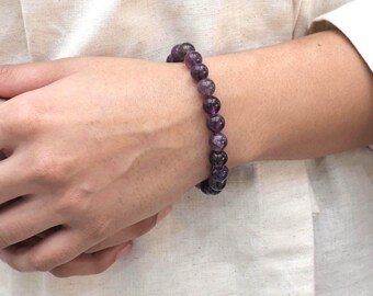 May include: A bracelet made of dark purple amethyst beads. The round, polished beads display various shades of purple. The bracelet is worn on a wrist, with the beads appearing to be approximately 0.8 cm in diameter.