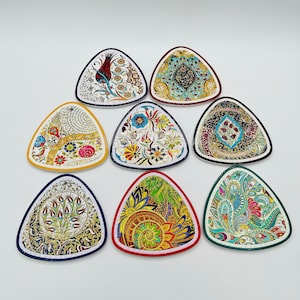 May include: Nine decorative, triangular ceramic plates with intricate floral and geometric patterns. Each plate features a unique design with a coloured border, including red, blue, and green. The plates are arranged on a white surface.