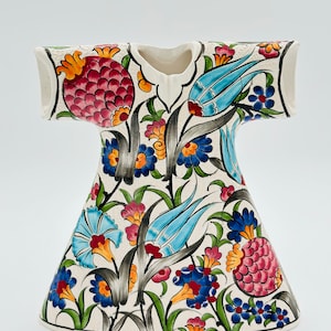 Puede incluir: Un jarrón de cerámica blanca con forma de vestido con un patrón floral en tonos de azul, rojo, verde, amarillo y negro.