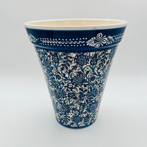 May include: A ceramic vase with a flared shape, featuring a detailed floral pattern in shades of blue and white. The rim is dark blue with a white dotted and swirl design. The interior is a cream color.