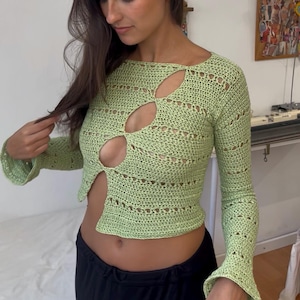 Può includere: Un top all'uncinetto verde chiaro a maniche lunghe con un design aperto unico. Il top presenta uno scollo tondo e maniche svasate. Il design aperto rivela uno scorcio dell'addome. Il top è abbinato a pantaloni neri.