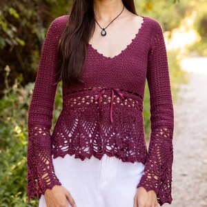 May include: A deep burgundy long-sleeve crochet top featuring a V-neckline, a fitted bodice with a thin ribbon tie at the waist, and flared, intricate lace-patterned sleeves. The garment is paired with plain white trousers and a dark pendant necklace.