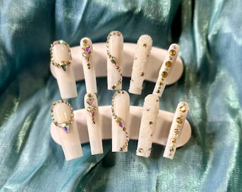 May include: A set of ten white press-on nails, each adorned with rhinestones and iridescent gems. Some nails feature a full rhinestone border, while others have a delicate rhinestone design. One nail is covered in small, white beads.