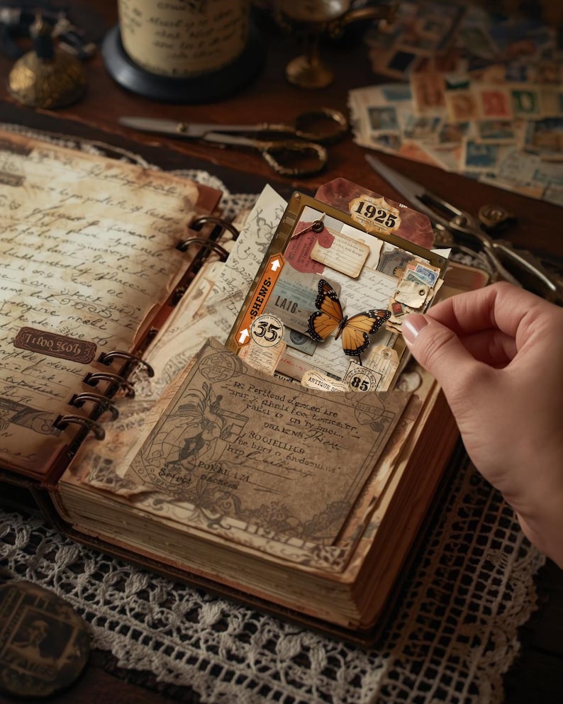 Puede incluir: Diario abierto con p&aacute;ginas de papel envejecido, con ef&iacute;meras vintage como pegatinas de mariposas, etiquetas y sellos. El diario est&aacute; encuadernado con una cubierta marr&oacute;n y anillos met&aacute;licos. Una mano sostiene una tarjeta con una mariposa.