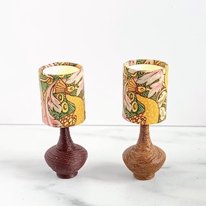 Op de afbeelding: Twee miniatuur tafellampen met patronen kappen. Eén lamp heeft een donkerrode voet, de andere een lichtbruine voet. De kappen hebben een bloemenpatroon met gele, roze en groene elementen. De lampen zijn decoratief.