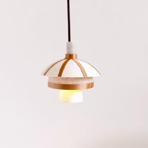 Op de afbeelding: Een kleine, moderne hanglamp met een wit en bruin gestreepte kap. Het licht hangt aan een zwart snoer en geeft een warme, gele gloed. De armatuur heeft een gelaagd ontwerp en staat tegen een witte achtergrond.