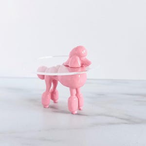 May include: A small, solid pink poodle figurine standing on four legs. The poodle has a smooth, glossy finish and features a clear, circular plastic collar or ring encircling its neck area, extending outward from the body of the dog sculpture.