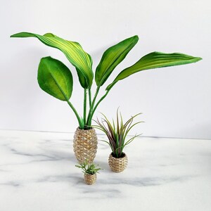 Op de afbeelding: Drie decoratieve kunstplanten in getextureerde goudkleurige potten. De grootste plant heeft brede groene bladeren, de middelste heeft dunne, stekelige bladeren en de kleinste heeft een compact, bladrijk uiterlijk. De potten hebben een ananasachtig ontwerp.