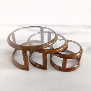 May include: Three nested glass and wood coffee tables. The tables are round and have a clear glass top with a brown wood frame.