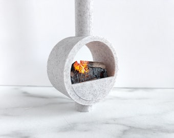 May include: A contemporary, grey speckled fireplace with a circular design. It has a small shelf holding a miniature fire with orange and yellow flames and a log. The fireplace is a decorative item, likely for a dollhouse or similar.
