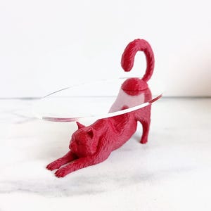 May include: A red cat figurine in a stretching pose, with a clear, oval-shaped object passing through its body. The cat is on a white surface, with a white background. The cat's tail is curled.