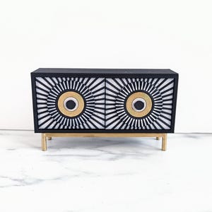May include: A black cabinet with two doors, featuring a geometric sunburst design in white and black. The cabinet has gold accents and sits on four gold-colored legs. A modern piece of furniture.