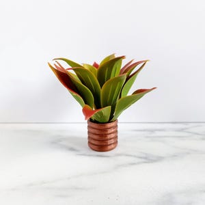 May include: A small artificial succulent plant with green and red-tipped leaves. The plant is in a small, brown, ribbed pot. The background is white.