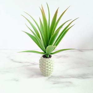 May include: A small, artificial succulent plant in a white, textured pot. The plant has long, green leaves with reddish-brown tips. The pot is shaped like a pineapple and sits on a white marble surface.