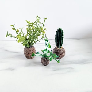 Op de afbeelding: Drie miniatuur kunstplanten in decoratieve bruine potten. De planten omvatten een groene cactus, een bladgroene plant en een kruipende groene plant. De potten hebben een textuur, geweven uiterlijk. De planten zijn gerangschikt op een wit oppervlak.