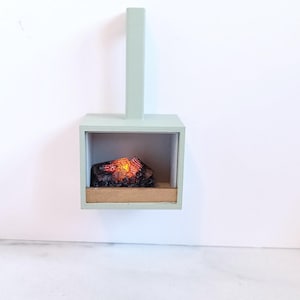 Op de afbeelding: Een miniatuur open haard in een lichtgroene tint, gemonteerd op een witte muur. De open haard heeft een rechthoekig doosontwerp met een gloeiend, gesimuleerd vuur erin. Onderaan een kleine, lichtbruine richel.