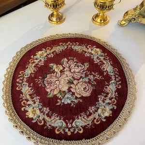 Könnte beinhalten: Eine runde bordeauxrote Stoffmatte mit einem zentralen Blumen-Gobelin-Design aus rosa und cremefarbenen Rosen, umgeben von einer kreisförmigen Bordüre aus bunten Blumen und Blättern, verziert mit einer beigen geflochtenen Borte am Rand.