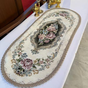 Può includere: Un runner da tavola ovale con base in tessuto color crema, caratterizzato da un medaglione centrale scuro e motivi floreali nei toni del rosa, blu e verde. Il runner è rifinito con un bordo decorativo in pizzo beige smerlato.