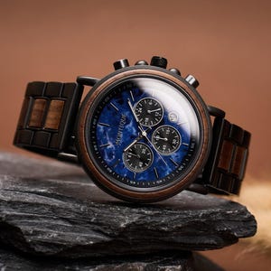 May include: A wooden wristwatch with a blue face and black subdials, featuring the brand name "MONTIQUE". The watch has a dark wood bezel and a matching wooden band. The watch is displayed on a dark gray rock.