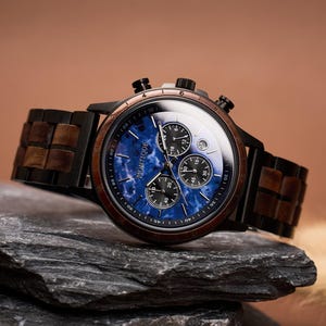 May include: A wristwatch with a blue face and a wooden band. The watch face has three sub-dials and the brand name "MONTIQUE" printed on it. The watch is set on a dark gray rock against a brown background.