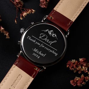May include: A black watch with a burgundy leather strap. The watch face is engraved with the word "Dad", the phrase "Thank you for everything", the name "Michael", and the year "2024". The watch has a silver-colored bezel.