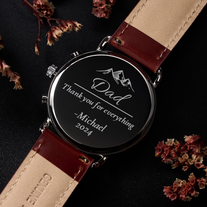 May include: A black watch with a burgundy leather strap. The watch face is engraved with the word "Dad", the phrase "Thank you for everything", the name "Michael", and the year "2024". The watch has a silver-colored bezel.