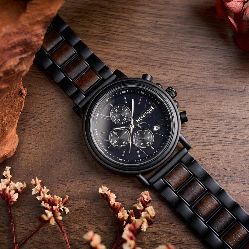 Op de afbeelding: Een zwarte Montique chronograaf horloge met een donkerblauwe wijzerplaat en een houten band. Het horloge heeft drie sub-wijzerplaten en een datumvenster. De band combineert zwarte metalen schakels met houten inzetstukken. Het horloge ligt op een houten oppervlak.