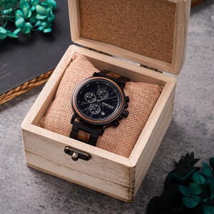 May include: A black and wood-effect wristwatch with a dark blue face, presented in an open wooden box. The watch has three sub-dials and the brand name "MONTIQUE". The box has a light brown interior and a small clasp.