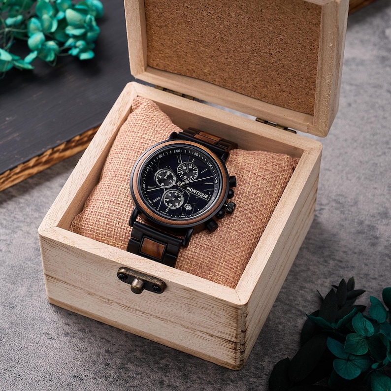 May include: A black and wood-effect wristwatch with a dark blue face, presented in an open wooden box. The watch has three sub-dials and the brand name "MONTIQUE". The box has a light brown interior and a small clasp.
