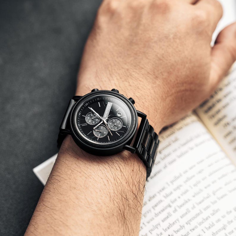 Op de afbeelding: Een zwart polshorloge met een ronde wijzerplaat en een zwarte metalen band wordt getoond op een pols. Het horloge heeft drie sub-wijzerplaten en een datumweergave. Het horloge staat tegen een donkere achtergrond.