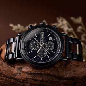 Op de afbeelding: Een zwarte Montique chronograaf horloge met een donkerblauwe wijzerplaat en houten accenten. Het horloge heeft meerdere sub-wijzerplaten en een datumweergave. Het horloge ligt op een houten oppervlak.