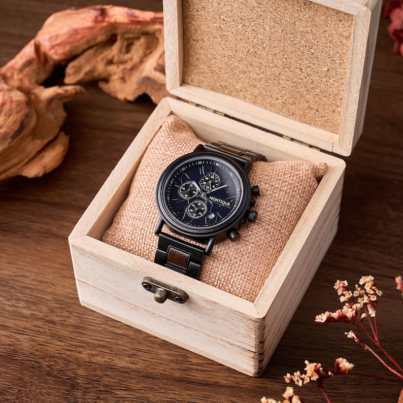Op de afbeelding: Een zwarte Montique chronograaf horloge met een donkerblauwe wijzerplaat, gepresenteerd in een open houten kist. De horlogeband is van zwart metaal met bruine houten details. De kist ligt op een beige kussen.