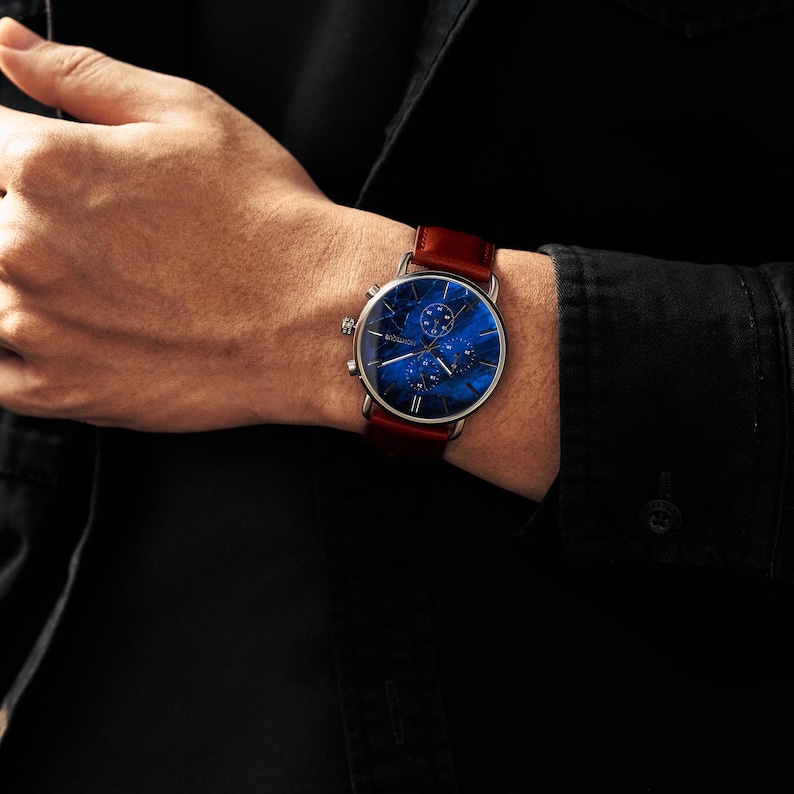 May include: A wristwatch with a dark blue face and a brown leather band. The watch face has a unique pattern with silver accents. The watch is on a wrist, with a black shirt in the background.
